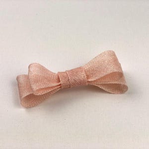 May include: A light pink bow made of a woven material. The bow is tied in the center and has two loops on each side. The bow is lying on a white surface.
