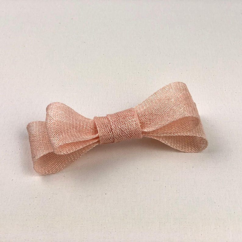 May include: A light pink bow made of a woven material. The bow is tied in the center and has two loops on each side. The bow is lying on a white surface.