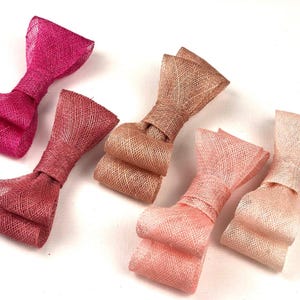 May include: Close-up of several fabric bow embellishments in shades of pink and peach. The bows are made of a woven material, with a textured appearance. The bows are tied in the center and have a classic bow shape. These bows are suitable for crafts and accessories.