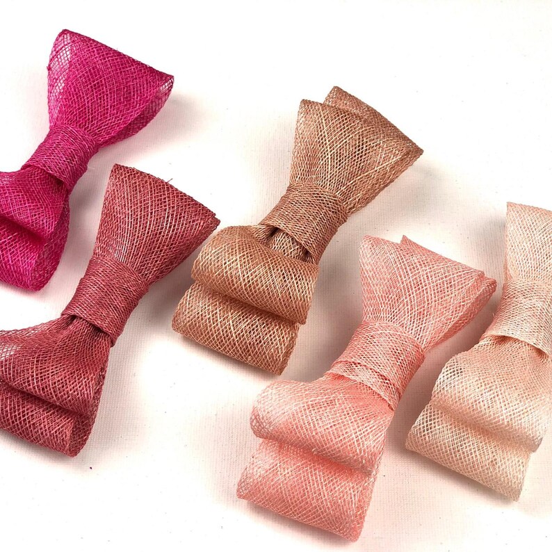 May include: Close-up of several fabric bow embellishments in shades of pink and peach. The bows are made of a woven material, with a textured appearance. The bows are tied in the center and have a classic bow shape. These bows are suitable for crafts and accessories.