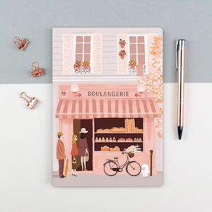 Puede incluir: Un cuaderno con una ilustración rosa de una panadería francesa, con la etiqueta "BOULANGERIE". La escena incluye personas, una bicicleta con una cesta y un perro blanco pequeño. Un bolígrafo y clips de carpeta de oro rosa están sobre la superficie gris y blanca.