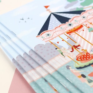 May include: A stack of blue and white notebooks with a colorful illustration of a carousel on the cover. The carousel has a pink and white striped roof, a blue and white striped canopy, and a red and white striped base. There are small flags and a red star on top of the carousel.