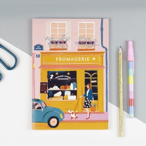 Può includere: Un quaderno illustrato rosa e giallo con una scena di facciata di negozio. La facciata è un negozio di formaggi chiamato "Fromagerie" con un cartello che dice "Fromages Affines Cremerie". Una donna con una giacca blu e una gonna a pois passeggia un cane al guinzaglio davanti al negozio. Un'auto blu è parcheggiata davanti al negozio.