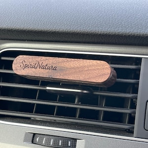 Könnte beinhalten: Ein hölzerner Auto-Luftauslassdiffusor mit dem Text "SpiritNatura" in kursiver Schrift. Der Diffusor ist rechteckig mit abgerundeten Kanten und wird in der Lüftungsöffnung eines Autos platziert. Das Armaturenbrett des Autos ist im Hintergrund sichtbar.