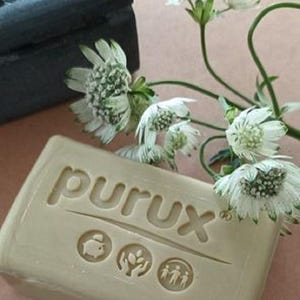 May include: A light-colored bar of soap with the word "purux" embossed on it, along with three small symbols. A dark gray box with a floral design is in the background, and white flowers with green accents are in the foreground.