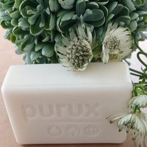 May include: A rectangular bar of white soap with the word "purux" embossed on the front. The soap is positioned in front of a backdrop of green foliage and white and green flowers.