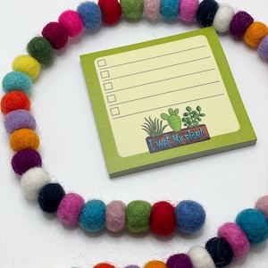 May include: Green checklist notepad with a yellow background and the text "I wet my plants". The notepad has a graphic of a potted cactus and other plants. The notepad is surrounded by a colourful garland of felt balls.