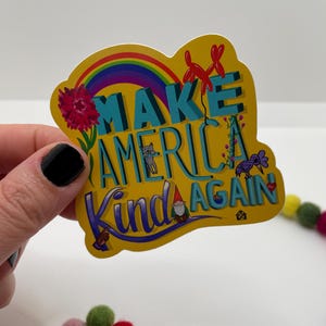 May include: Yellow sticker with a rainbow, a gnome, a cat, and birds. The sticker says "Make America Kind Again."