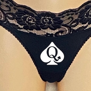 Könnte beinhalten: Schwarze Unterwäsche mit einem weißen Pik-Symbol mit einem stilisierten "Q"-Design. Der obere Rand ist mit schwarzer floraler Spitze verziert. Das Kleidungsstück ist aus einem weichen, dunklen Stoff gefertigt.