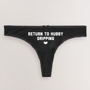Puede incluir: Braga tanga negra con el texto blanco "RETURN TO HUBBY DRIPPING" y un corazón blanco. Los bordes están adornados con encaje y un pequeño lazo en la parte superior. Un diseño divertido.