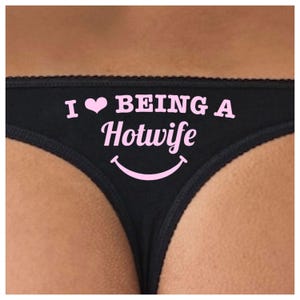 Op de afbeelding: Zwarte string met de roze tekst "I ♥ BEING A Hotwife", een roze hart en een smiley. De string heeft een delicate kanten rand.
