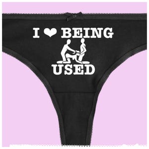Könnte beinhalten: Schwarzer Tanga mit einem weißen grafischen Aufdruck, der "I ♥ BEING USED" mit einer Strichmännchen-Illustration sagt.