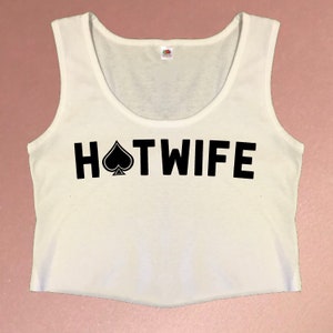 May include: White crop top with a scoop neck and the text "HOTWIFE" in bold black letters, with a black spade symbol replacing the "O". The top is sleeveless and made of a soft material.