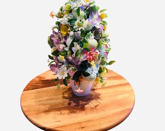 May include: A white ceramic planter filled with a variety of pastel colored Easter eggs and artificial flowers, including purple, pink, blue, and white flowers. The planter is sitting on a wooden round base.