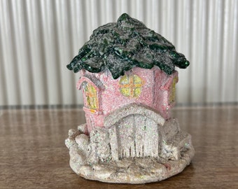 May include: A small pink and white gingerbread house with a green roof and a snowy base. The house has a door and two windows.
