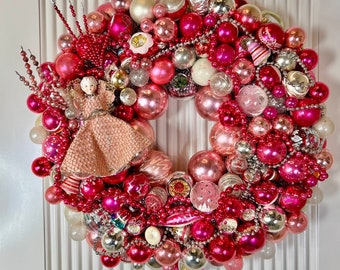 Op de afbeelding: Een roze en zilveren kerstkrans met een verscheidenheid aan ornamenten, kralen en een kleine porseleinen pop in het midden.