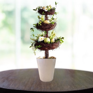 May include: A tiered Easter centerpiece with three nests filled with faux eggs and white flowers. The nests are stacked on a wooden pole and placed in a white ceramic pot.