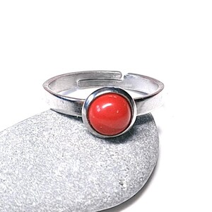 May include: A silver adjustable ring featuring a vibrant red, round stone. The ring sits on a grey stone, set against a white backdrop. The ring's band is made of polished metal.