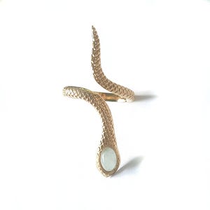 May include: A gold-toned metal ring shaped like a coiled snake with a textured, scale-like pattern along the band. The head of the snake features a smooth, oval-shaped, pale green stone inlay. The ring has an open, adjustable design.