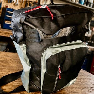 Könnte beinhalten: Ein schwarzer Rucksack mit einer großen Vordertasche mit hellgrauem Rand, einem Tragegriff und schwarzen Schultergurten. Der Rucksack verfügt über silberne Reißverschlüsse mit roten Kordelzügen, ein horizontales Netzteil und ein kleines Logo.