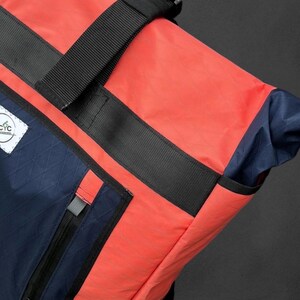May include: A close-up of a backpack with a vibrant color scheme. The bag features a combination of coral, navy blue, and black. The design includes a black strap with a buckle, a zippered pocket, and a small white label with text.