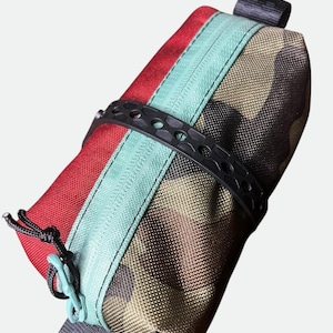Könnte beinhalten: Eine rechteckige Reißverschlusstasche mit einem roten Bereich und einem camouflage-gemusterten Bereich in Braun-, Grün- und Schwarztönen. Die Tasche hat einen hellblauen Reißverschluss, ein schwarzes, perforiertes Gummiband um die Mitte und schwarze Schlaufen.