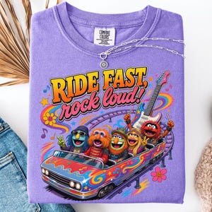May include: A purple t-shirt featuring a graphic of Muppet characters riding a colorful roller coaster car. The text "RIDE FAST, rock loud!" is printed above the characters. A guitar, musical notes, and flowers are included in the design.