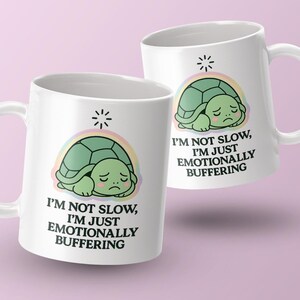 May include: Two white ceramic mugs with a cartoon turtle design. The turtle is green and sad, set against a rainbow and a starburst. Text on the mugs reads "I'M NOT SLOW, I'M JUST EMOTIONALLY BUFFERING."