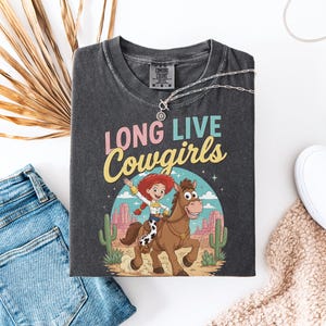 May include: A charcoal gray t-shirt featuring the text LONG LIVE Cowgirls in pink and yellow lettering. The graphic depicts a red-haired character riding a brown horse in a desert landscape with cacti, mesas, and a blue sky with white clouds.