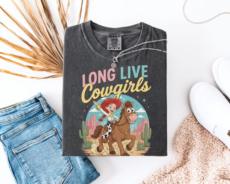 May include: A charcoal gray t-shirt featuring the text LONG LIVE Cowgirls in pink and yellow lettering. The graphic depicts a red-haired character riding a brown horse in a desert landscape with cacti, mesas, and a blue sky with white clouds.