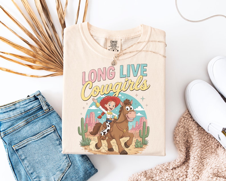May include: A cream-colored short-sleeve t-shirt featuring a graphic print of a cowgirl riding a brown horse in a desert landscape. The text "LONG LIVE Cowgirls" is printed above the illustration in pink and yellow lettering.