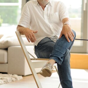 May include: A man wearing a white shirt, blue jeans, and brown shoes sits on a white folding chair. The chair is made of wood and has a white finish.