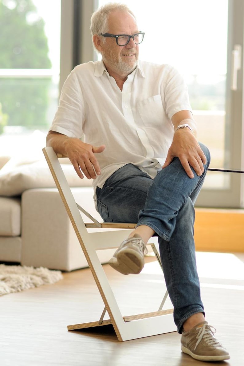 May include: A man wearing a white shirt, blue jeans, and brown shoes sits on a white folding chair. The chair is made of wood and has a white finish.