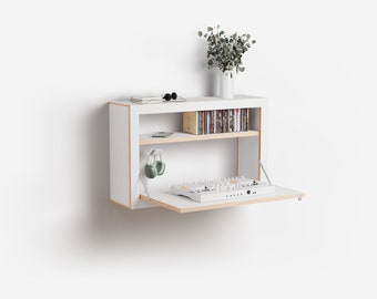 May include: A white wall-mounted shelf with a fold-down front. The shelf has two shelves, one with a row of CDs and the other with a pair of headphones. The front of the shelf is folded down to reveal a white DJ mixer.
