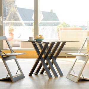 May include: A glass-top table with a unique criss-cross wooden base, surrounded by two folding chairs. One chair is gray and the other is white.