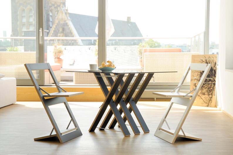 May include: A glass-top table with a unique criss-cross wooden base, surrounded by two folding chairs. One chair is gray and the other is white.