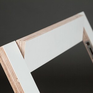 May include: Close-up of a white painted wooden frame with visible layers of plywood. The frame is angled and appears to be part of a larger piece of furniture.