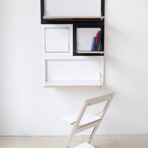 May include: A white and black wall-mounted desk with shelves and a fold-down desk surface. A white folding chair is in front of the desk.