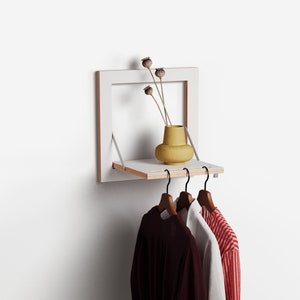 Op de afbeelding: Een witte wandplank met een ingebouwde kledingrek. Een gele vaas met gedroogde bloemen staat op de plank. Drie houten kleerhangers houden kleding vast. De plank heeft een natuurlijke houten rand.