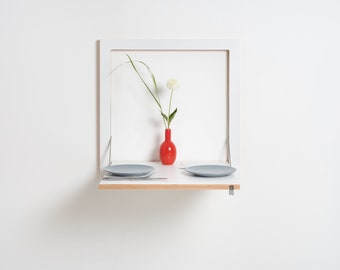 May include: A white wall-mounted folding table with a wooden top and two gray plates. A red vase with a white flower is on the table.