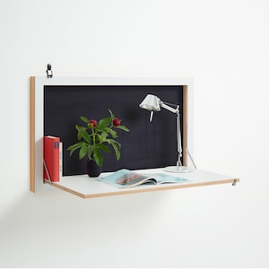 May include: White wall-mounted desk with a fold-down surface. The desk has a black interior and a silver lamp with a white shade. A book and a magazine are on the desk.
