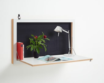 May include: White wall-mounted desk with a fold-down surface. The desk has a black interior and a silver lamp with a white shade. A book and a magazine are on the desk.