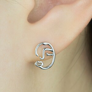 May include: A silver-colored stud earring in the shape of a minimalist face profile is shown. The earring is attached to an ear. The face design includes a curved line for the nose, a closed eye, and a pair of lips. The brand name "Bambi & dew" is visible.
