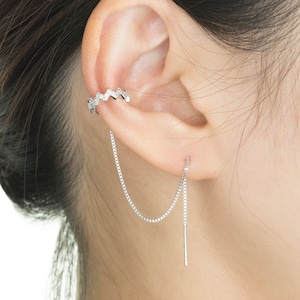 May include: A silver ear cuff with a zigzag design and a dangling chain earring. The ear cuff is adorned with small, sparkling stones. The earring features a delicate chain that connects to a straight bar. The jewelry is worn on the ear.