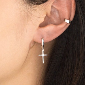May include: A silver hoop earring with a cross pendant hanging from it. A silver ear cuff is also visible. The jewellery is shown against a backdrop of dark hair and skin.