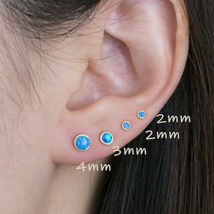 Peut inclure: Gros plan sur une oreille ornée de quatre petites boucles d'oreilles à clous rondes en opale bleue, serties d'or. Les boucles d'oreilles mesurent 2 mm, 2 mm, 3 mm et 4 mm. Le texte indique la taille de chaque boucle.