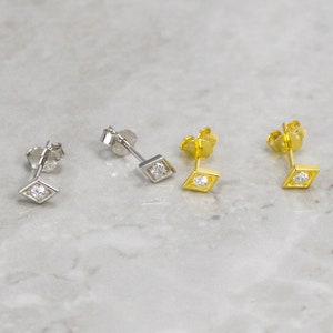May include: Four stud earrings with a diamond-shaped design. Two are silver-colored, and two are gold-colored. Each earring features a small, clear gemstone set in the center of the diamond shape. The earrings are displayed on a light gray marble surface.