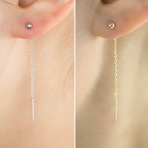 May include: A pair of earrings, one silver and one gold, each with a small ball stud and a fine chain that hangs down. The earrings are displayed on a person's ear.