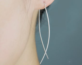 May include: Close-up of a silver earring with a curved, elongated design. The earring is suspended from a small chain and crosses at the bottom. The jewelry is shown against a muted teal background.