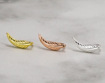 May include: Three feather-shaped ear cuffs in gold, rose gold, and silver. Each cuff features a detailed vein design and a curved shape, suggesting they are designed to wrap around the ear. The jewelry pieces are displayed on a light gray surface.